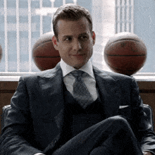 a man in a suit and tie sits in front of a window with basketballs in the background