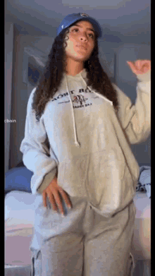 a woman wearing a hoodie and a blue hat is standing in a bedroom .