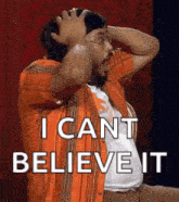 a man in an orange shirt is holding his head and says i can 't believe it .