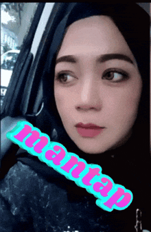 a woman wearing a hijab is sitting in a car and the word mantap is on the side of her face