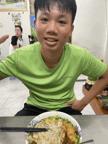 a young man wearing a green john henry shirt sits at a table with a bowl of food