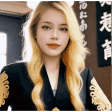 a woman with blonde hair and blue eyes is wearing a black jacket with gold embroidery on the sleeves .