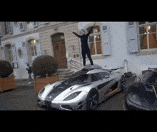 a woman stands on top of a silver sports car in front of a building