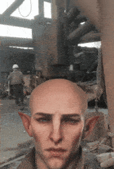 a bald man with elf ears looks at the camera with a man in a hard hat in the background