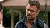a man with a beard is wearing a plaid shirt and a vest and talking to another man .
