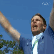a man in a blue shirt and tie is raising his fist in the air with the olympic rings in the background