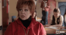a woman in a red jacket is sitting at a desk in a classroom with a group of children .