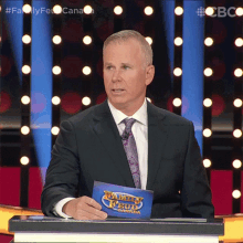 a man in a suit and tie stands at a podium holding a card that says family feud canada