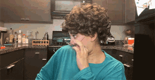 a person with curly hair covering their nose in a kitchen with a bottle of vodka in the background