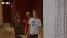 a man in a white t-shirt is walking down a hallway with a tabii logo in the background