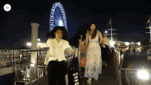 two women are walking across a bridge with a ferris wheel in the background and a sign that says i am on it