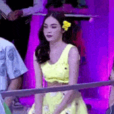 a woman in a yellow dress with a flower in her hair is standing in front of a purple wall .