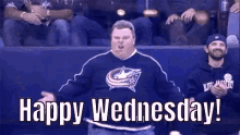 a man in a blue sweater is standing in front of a crowd of people and says `` happy wednesday ! ''