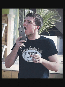 a man wearing a black shirt that says clermont lounge is holding a cell phone in his mouth