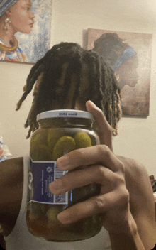 a person with dreadlocks holds a jar of pickles in front of their head