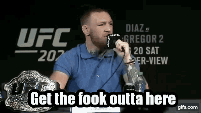 a man in a blue shirt is talking into a microphone with the words get the fook outta here below him