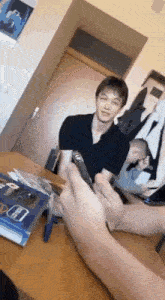 a man is sitting at a table holding a cell phone while another man holds a remote control .