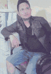 a man wearing a black shirt and a leather jacket sits in a chair