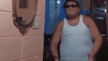 a man wearing sunglasses and a white tank top is standing in front of a mirror in a bathroom .
