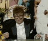 a man in a tuxedo and bow tie is sitting at a table in front of a screen that says volver