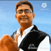 a man wearing glasses and a black vest is smiling in front of a blue sky
