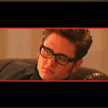 a man wearing glasses is sitting on a couch and looking at the camera .