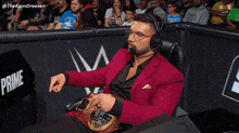 a man in a red suit is sitting in a chair with a microphone on his head and a championship belt around his waist