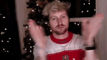 a man with a beard wearing a christmas sweater is standing in front of a christmas tree with his hands up .