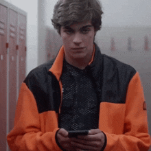a young man in an orange and black jacket is looking at his cell phone