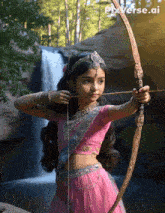 a girl in a pink dress is holding a bow and arrow in front of a waterfall with pixverse.ai in the corner