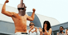 a shirtless man flexes his muscles in front of a group of people including a woman wearing sunglasses