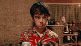 a young man wearing a hawaiian shirt is sitting on a couch holding a can of soda .
