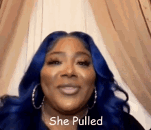 a woman with blue hair and hoop earrings is standing in front of a curtain and smiling .