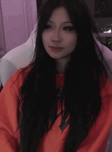 a woman with long black hair is sitting in a chair wearing an orange sweatshirt .