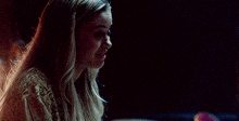 a woman with long blonde hair is sitting in a dark room looking down at something .