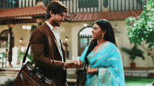 a man in a suit and a woman in a blue saree are shaking hands in front of a building .
