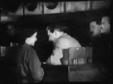 a black and white photo of a group of people standing around a table in a dark room .
