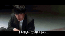 a man in a suit is sitting on a bed with korean writing on the screen behind him .