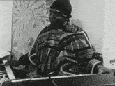 a black and white photo of a man in a poncho playing a piano .