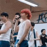a man with red hair is singing into a microphone while a group of people are dancing in a gym .
