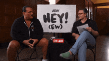 two men sitting in front of a sign that says hey