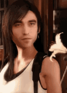 a man with long hair and a white tank top is standing next to a flower .