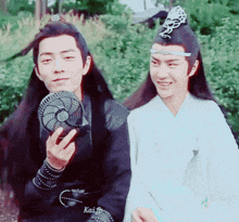 two men are sitting next to each other and one of them is holding a fan in his hand .