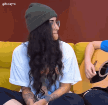 a woman wearing a beanie and glasses is sitting next to a man playing a guitar ..