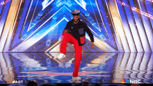 a man in red pants and a hat is dancing on a stage with a nbc logo behind him