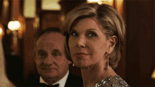 a woman in a silver dress looks at the camera while a man in a tuxedo sits behind her