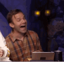 a man in a plaid shirt is laughing while sitting in front of a tablet computer .