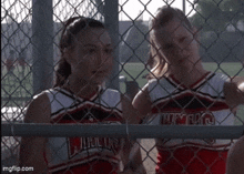 two cheerleaders are behind a chain link fence and one is wearing a uniform that says ' lions ' on it