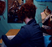 a woman with red hair sits at a table in front of a painting