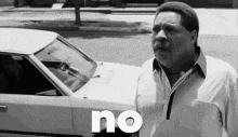 a man in a white shirt is standing in front of a white car and saying no .